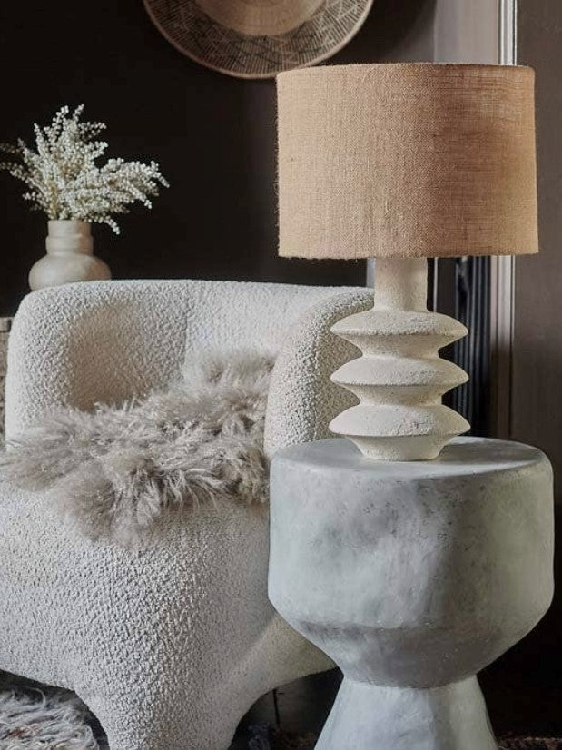 The Akito lamp with a creamy hued textured base and a jute lampshade, placed next to a white chair with a fluffy blanket on it, in a cozy home setting.