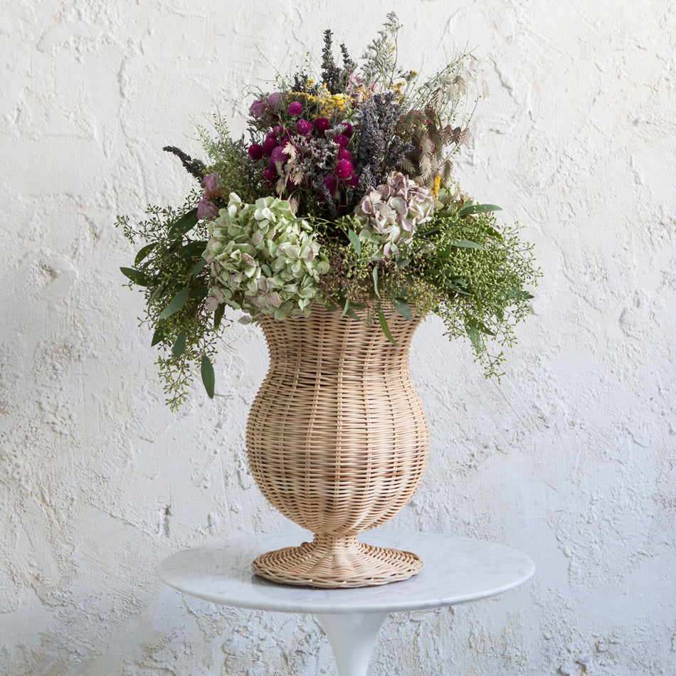 Rattan urn with a bouquet of flowers on a white pedestal against a textured white background