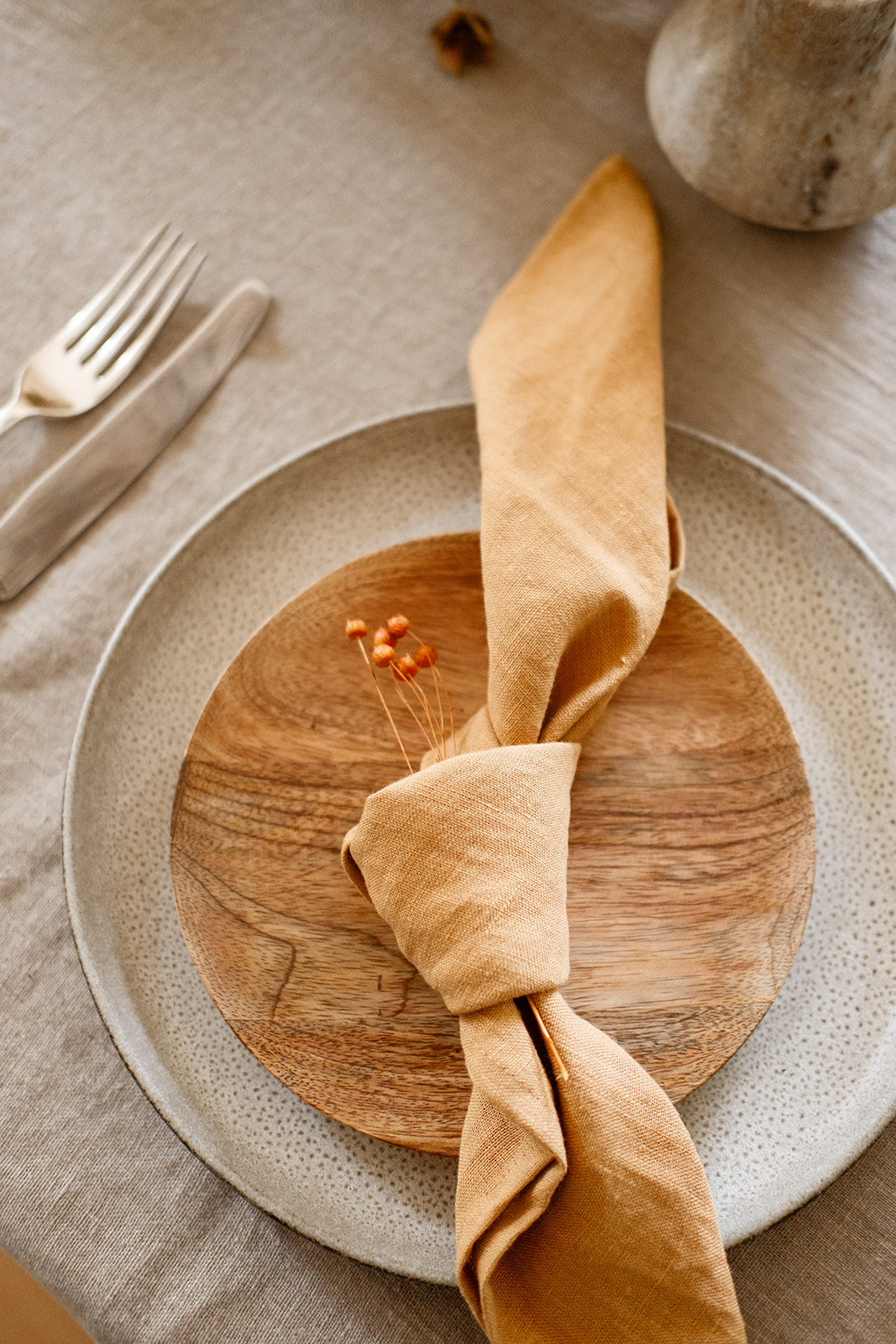 Detail shot of a place setting with natural linen tablecloth, textured grey stoneware dinner plate, wood side plate with Cinnamon coloured stonewashed linen napkin tied in a knot and laid on top.