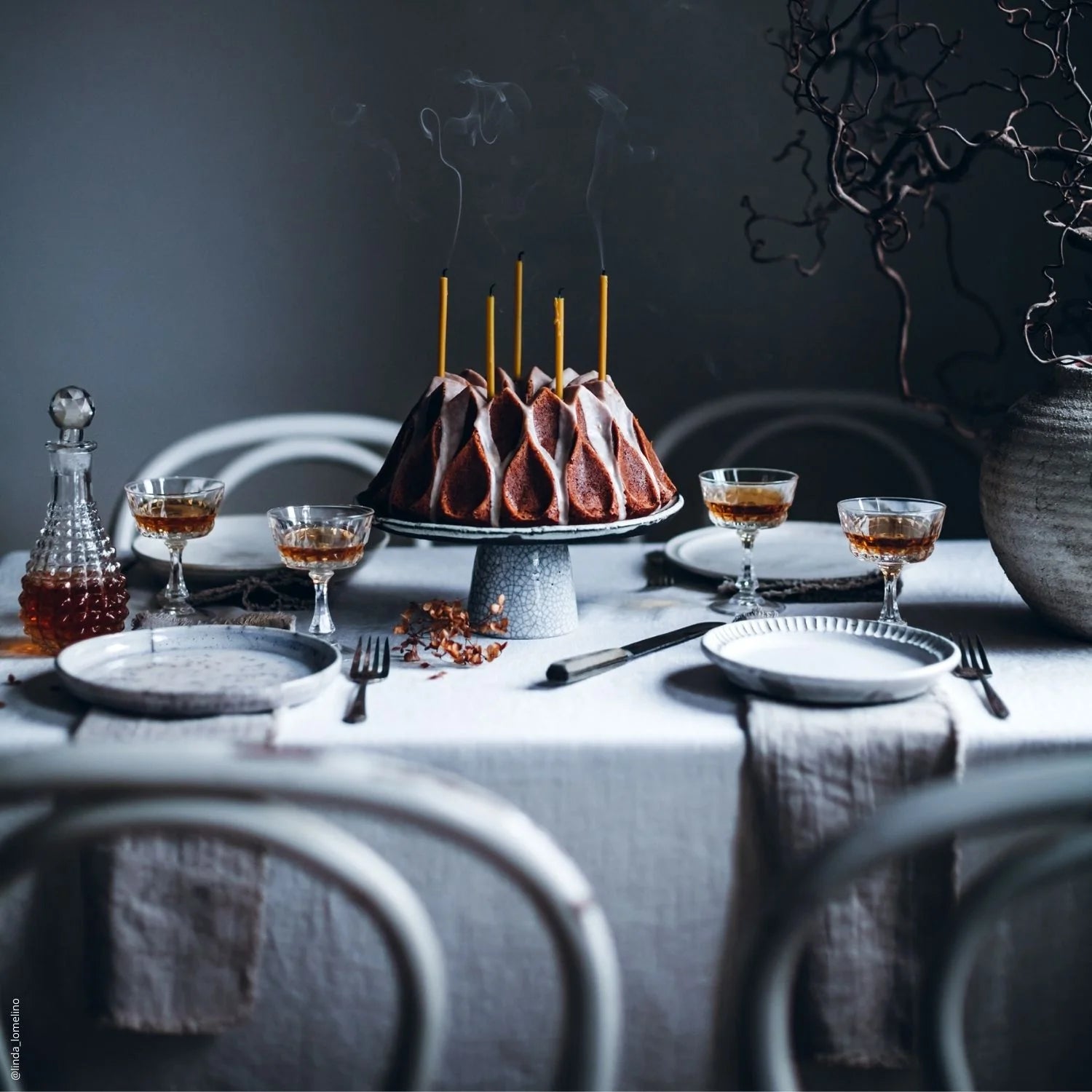 View of a tablescape with moody grey and beige colour palette; vase with twisted willow branches, a cake on top of a cake stand with ochre candles, 4 place settings with natural linen napkins with fringes and textured white plates and glassware.