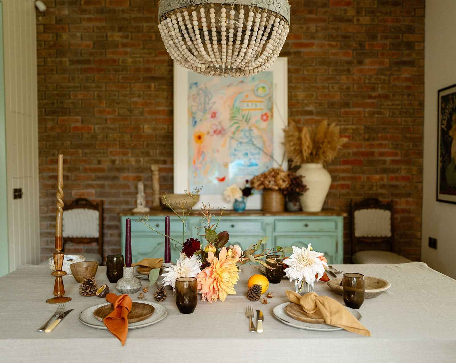 Dining room with a table set for a meal, featuring a wooden bead chandelier, brick wall, and decorative elements.
