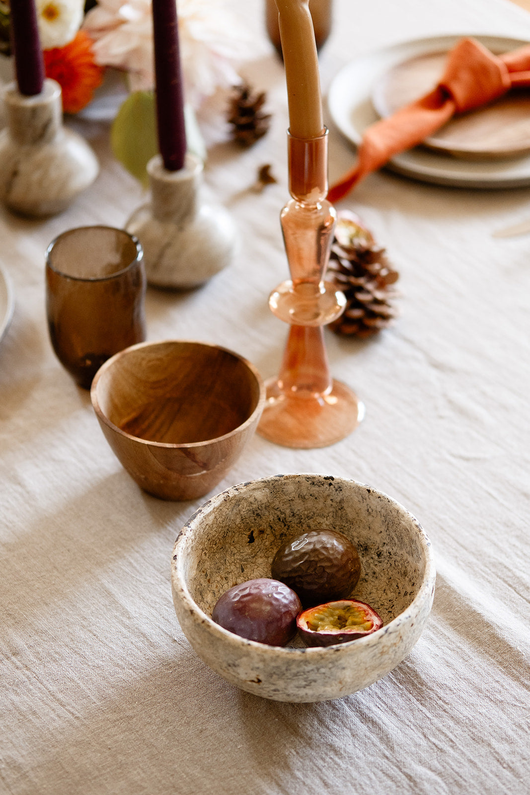 Close up of Earthy table setting with wooden bowls, rustic cream bowl, rust glass candle holder, marble candles and pine cones on a natural linen tablecloth