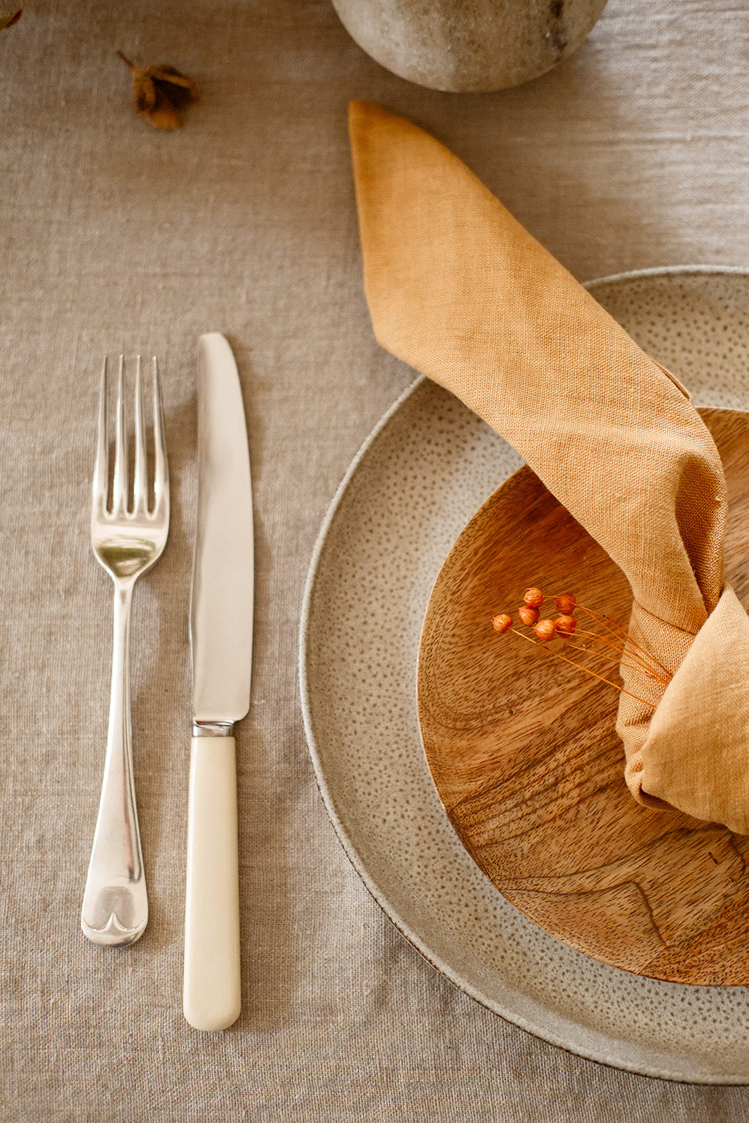 Detail shot of a place setting with textured grey plate with wood side plate on top and a cinnamon coloured linen napkin knotted on top of it with vintage cutlery on a natural linen tablecloth