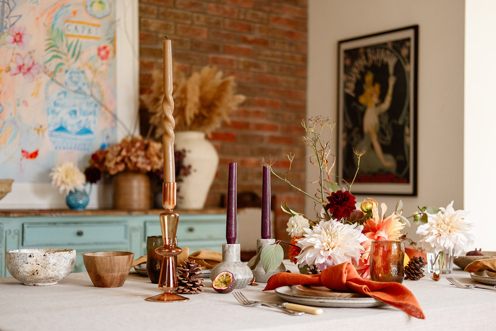 Lifestyle shot of an earthy tablescape with earthy terracottas, rust, brown, orange tones with natural linen tablecloth, rust glass candle holder with tall twisty brown candle, wooden bowl, marble candle holder with plum candle, pine cones, cream dahlias with brick wall and colourful painting in the background.