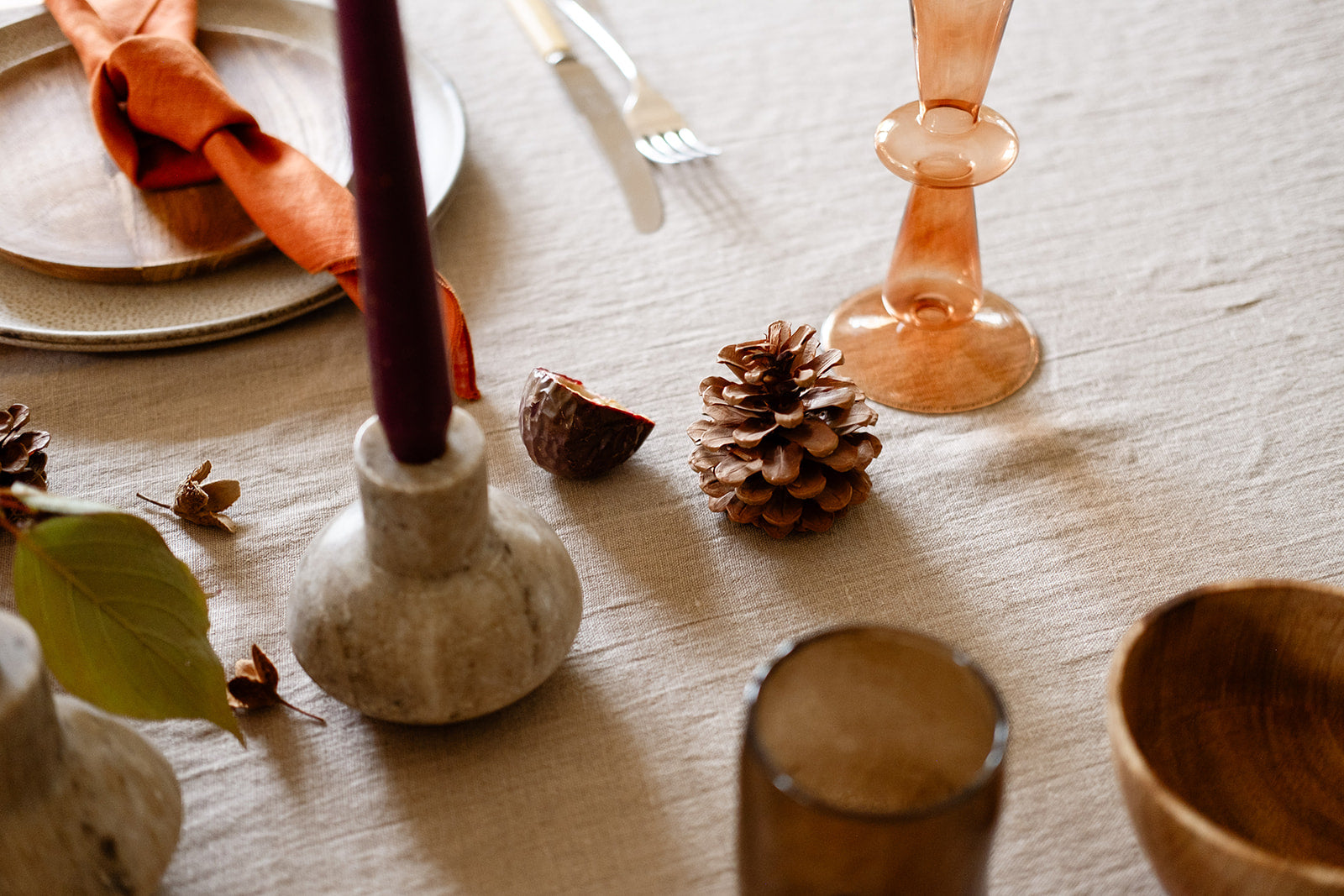 Detail shot of table setting with stonewashed natural linen tablecloth, marble candle holder with plum candle, rust glass candle holder, pine cones, figs, terracotta napkin, brown glass, wooden bowl