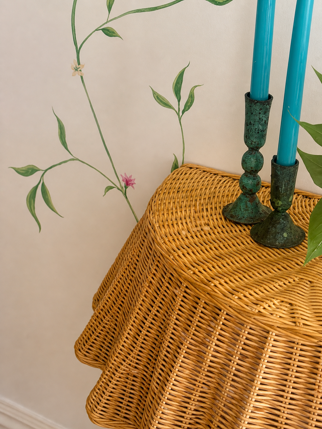 Details of rattan skirted detail of floating rattan table against hand painted botanical mural with verdigris candlesticks on top