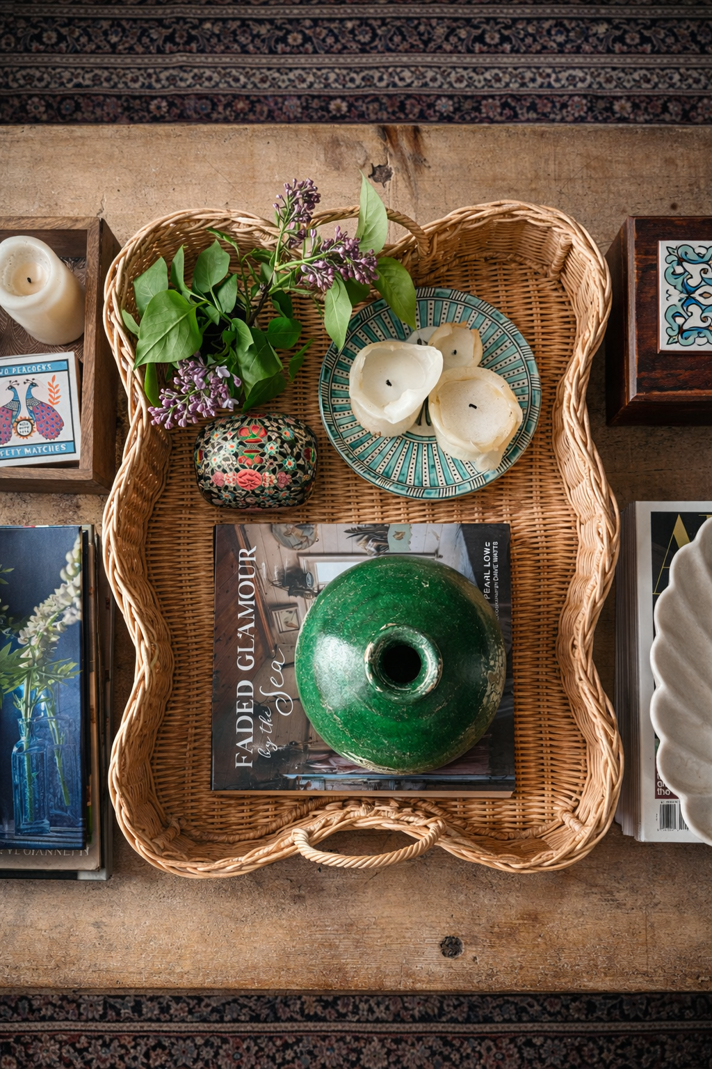 Overhead view of scalloped rattan tray on rustic coffee table with books, candles, decorative trinket box, round green vase, decorative bowls, fresh flowers