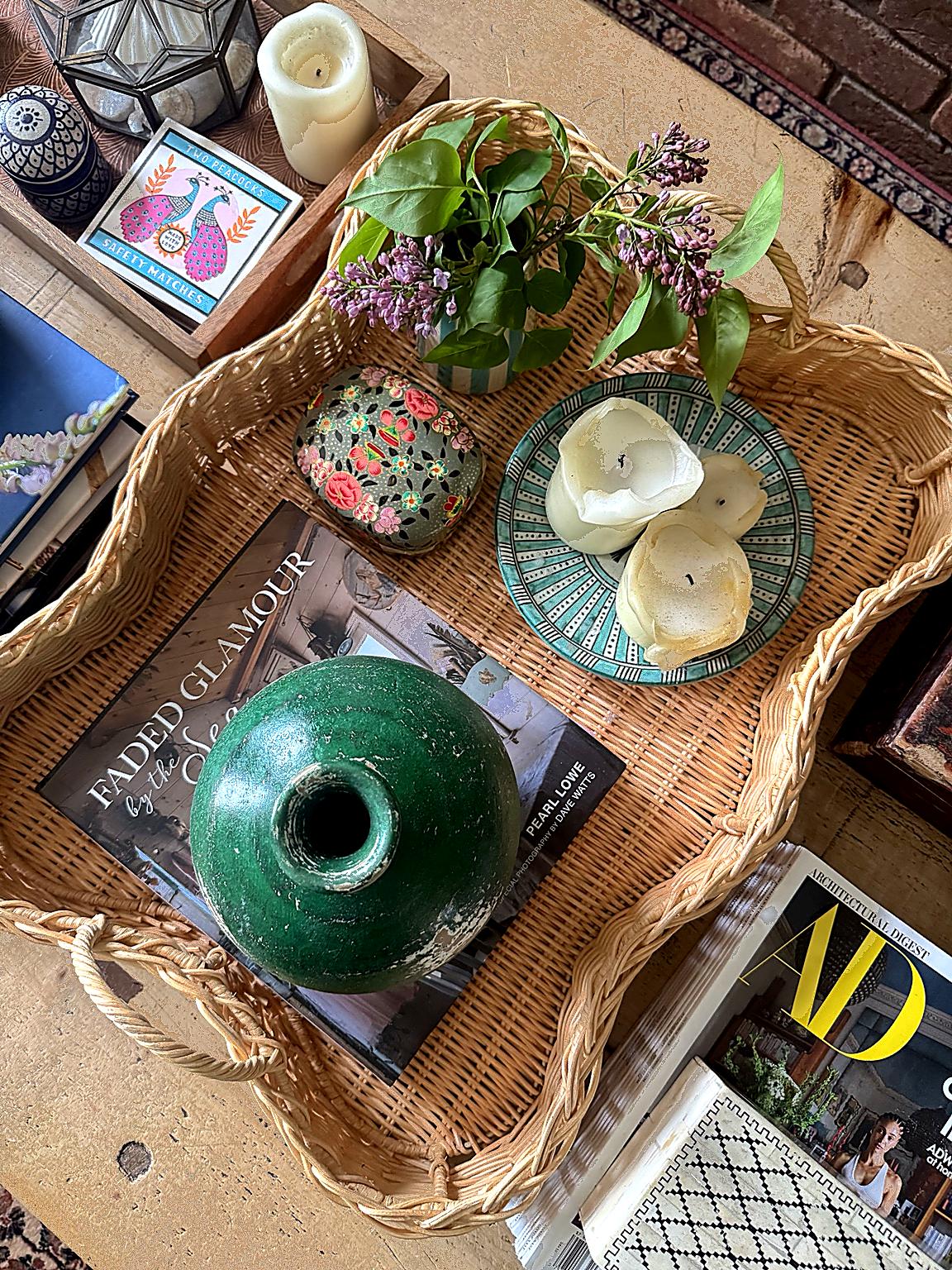 Overhead view of scalloped rattan tray on rustic coffee table with books, candles, decorative trinket box, round green vase, decorative bowls, fresh flowers