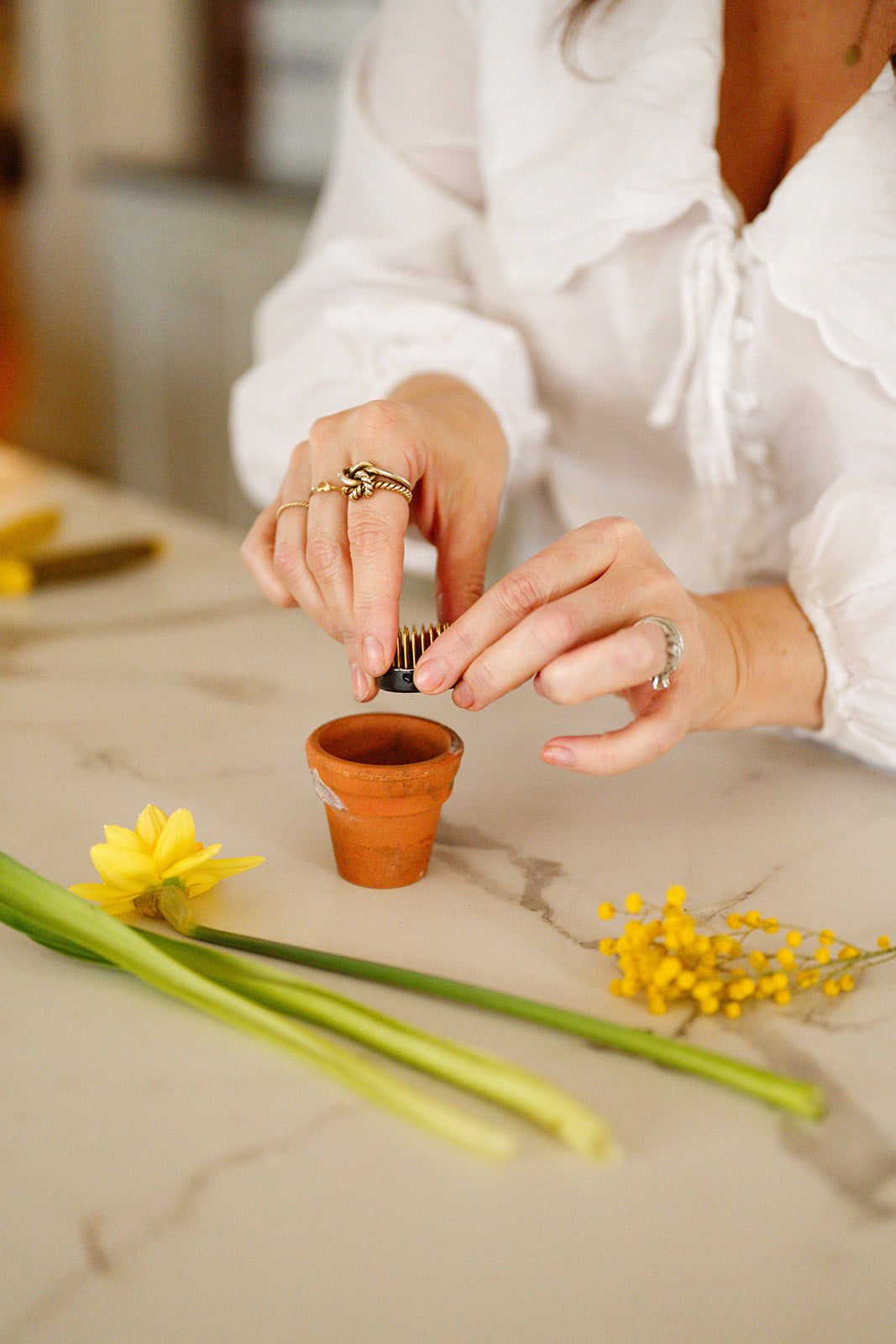 Woman placing small flower frog into mini terracotta pot on white marble counter with spring flowers laid out