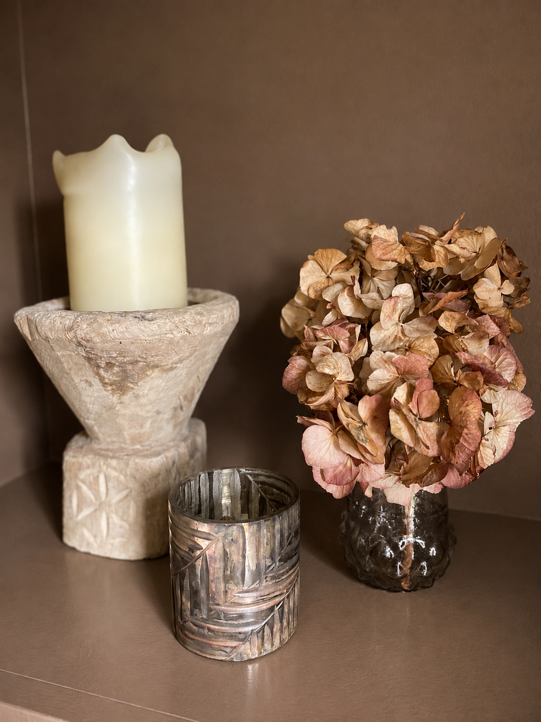A styled interior scene with a cream candle in a textured stone holder, a small metallic candle holder, and a vase of dried pink and tan hydrangeas against a warm brown wall and shelf.