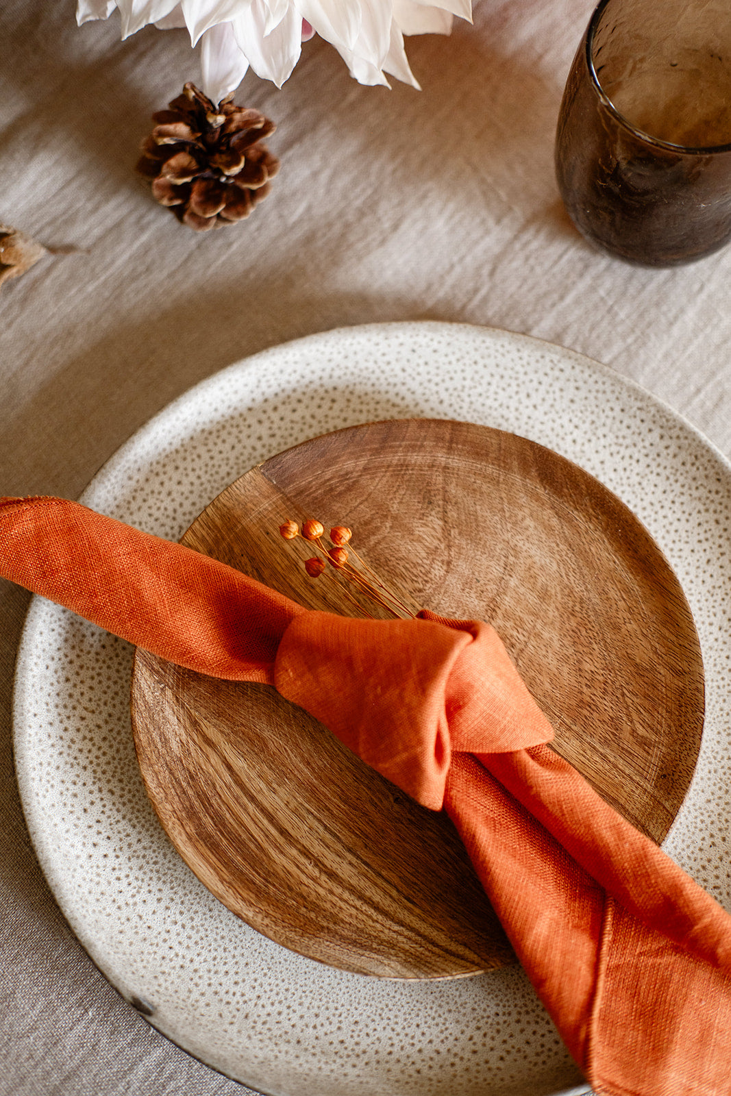 Overhead view of place setting with a knotted terracotta linen napkin sitting on a wood plate on top of a textured gray plate on a linen tableclot with brown glass