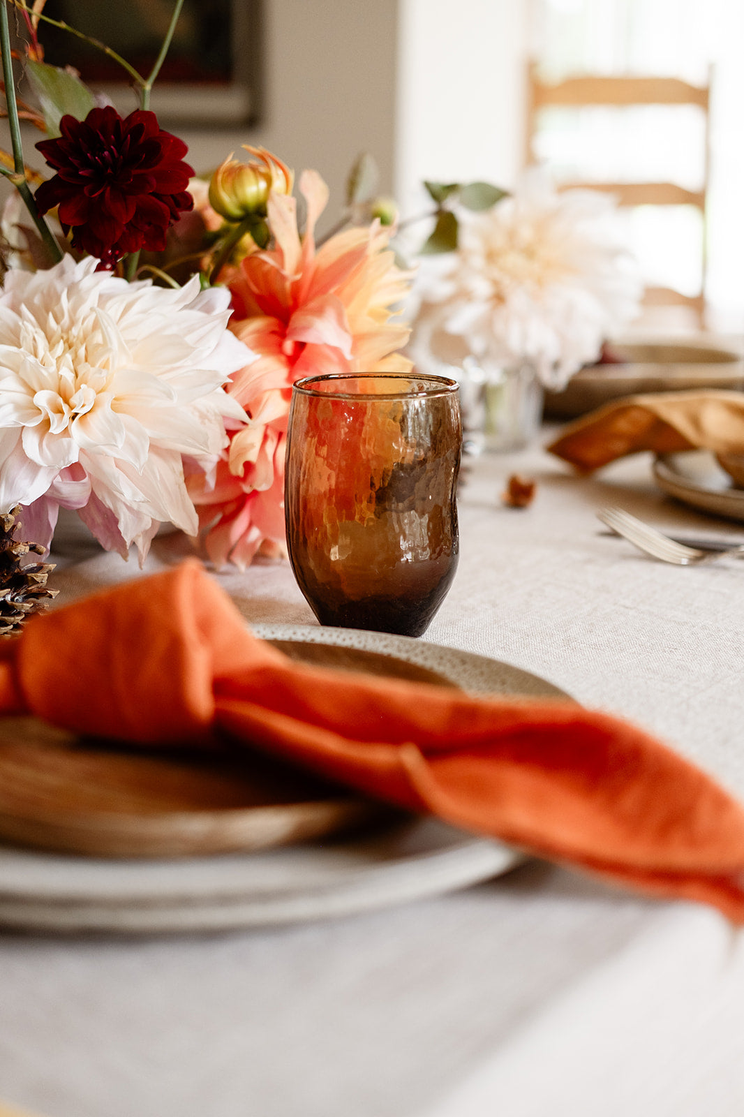 terracotta napkin arranged on place setting with wood plate and textured plate with brown drinking glass and cream and orange flowers