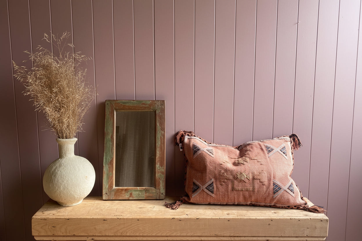 Wide shot of reclaimed wood mirror with weathered green finish on wooden bench against dusky pink tongue and groove wall next to vase of pampas and bohemian cushion
