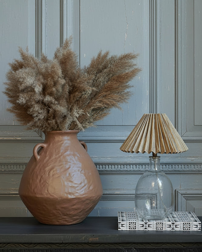 Pampas grass in brown textured vessel next to a small glass lamp with pleated shade against old panelled wall