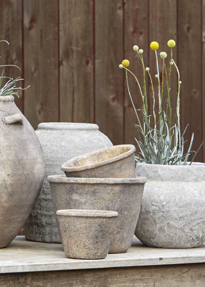 A collection of aged brown concrete pots of various sizes displayed outdoors, some with plants growing in them.