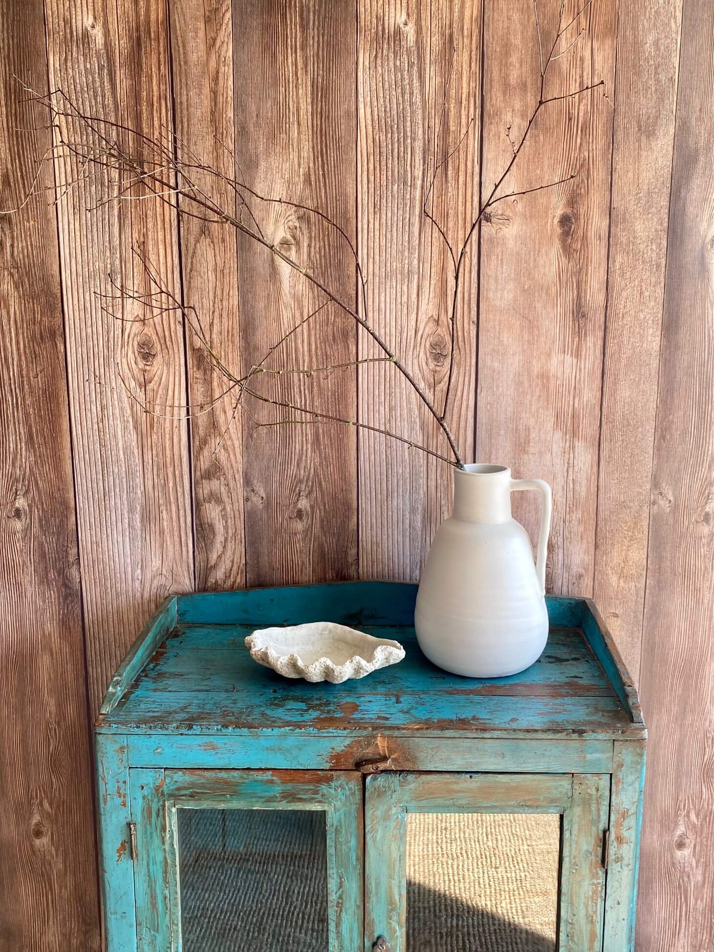 Textured cement bowl styled next to matte white glazed pitcher vase with sculpturual branches sitting on a vintage Indian turquoise cabinet with glass doors against a rustic wood plank wall.