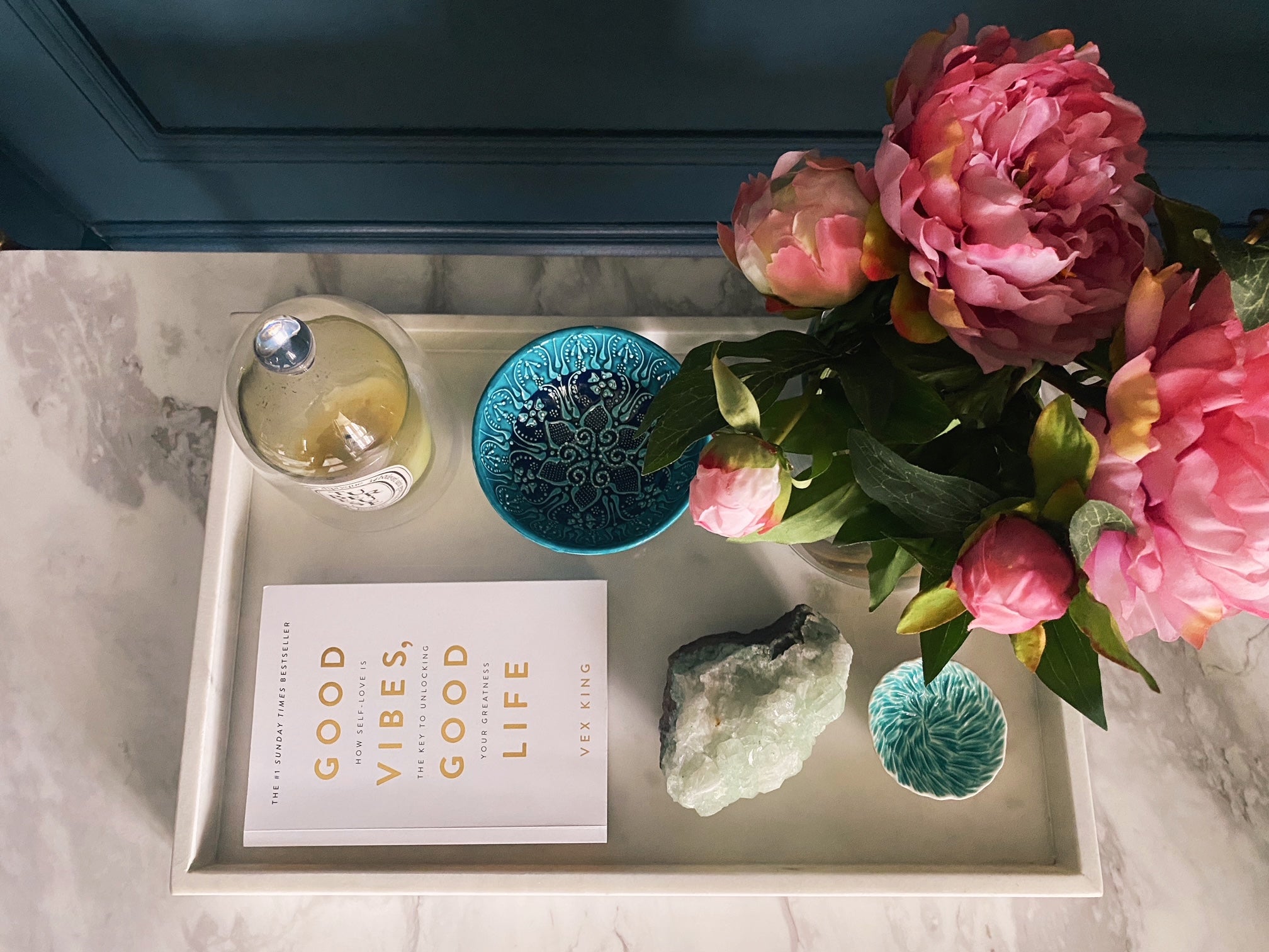 Overhead view of white marble tray with pink flowers, candles, books and mixture of turquoise trinket bowls