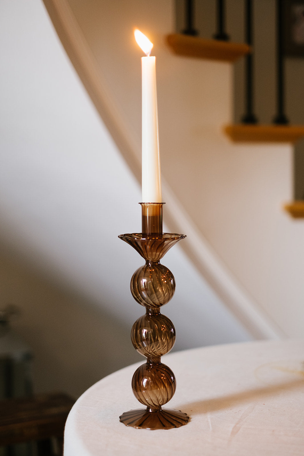 Brown glass candleholder with cream candle lighting in a cream room with black stair balustrade in background