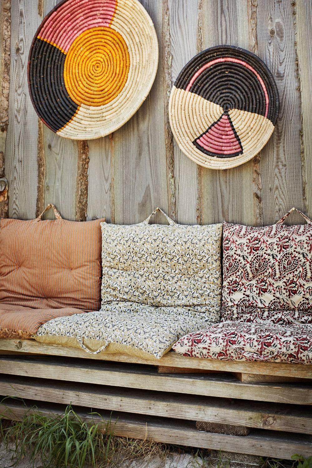 The image shows two circular woven plates with geometric patterns in red, yellow, and black, hanging on a wooden fence. Below, there is a rustic pallet bench with bohemian block printed patterned mattresses in different colours and prints.
