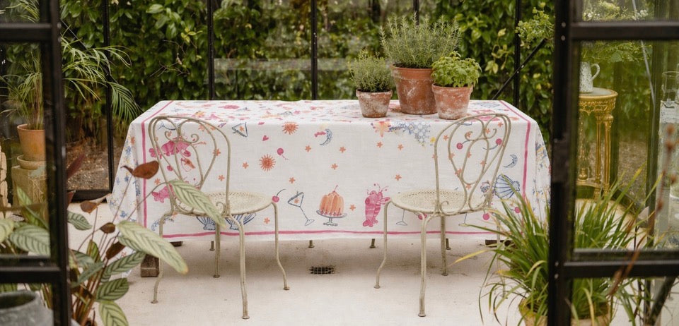 Hand painted linen tablecloth on table in glass house setting surrounded by greenery 