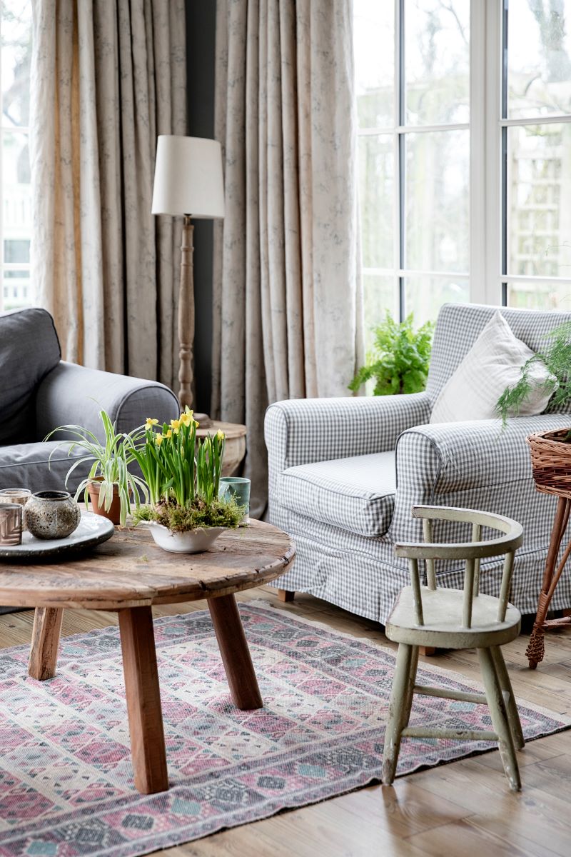 Round reclaimed wood coffee table in cosy living room with large windows, linen curtains, gingham armchair and wooden lamp in corner
