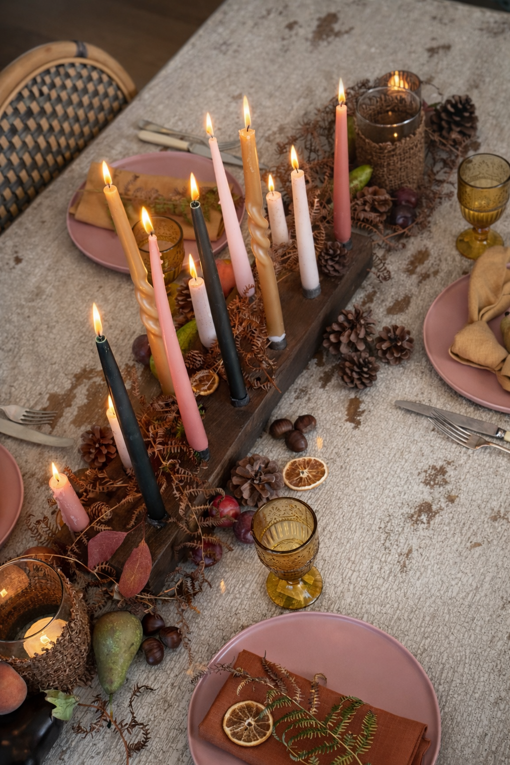 Decorative table setting with candles, pinecones, and pink plates on a textured surface.