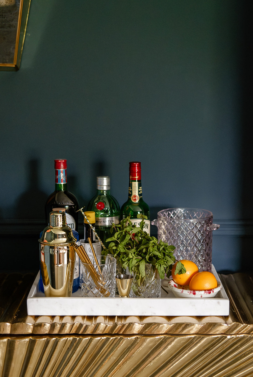 Front view of a cocktail tray of spirits, rose-hued glass ice bucket, gold cocktail maker, mint and gold straws on an art deco style gold cabinet in moody living room