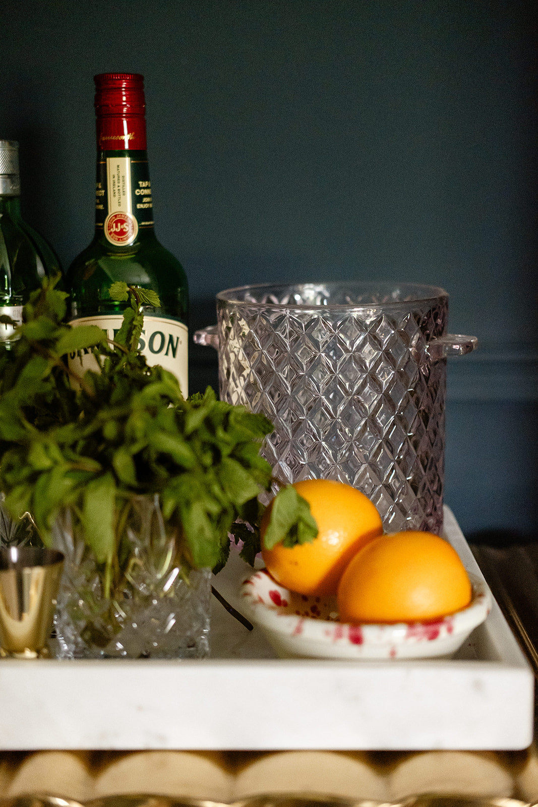 Rose ice bucket on white cocktail marble tray next to spirits bottles, bowl of oranges and glass of mint