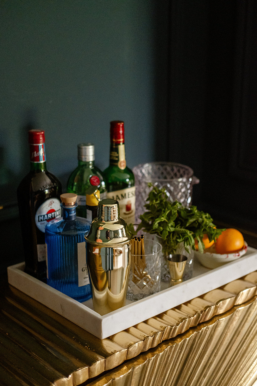 Bar tray with cocktail ingredients including bottles, a shaker, and fruits on a dark background