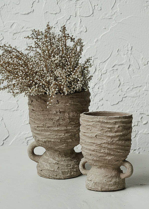 Browny beige raw concrete textural vase in hourglass shape  against textured cream background