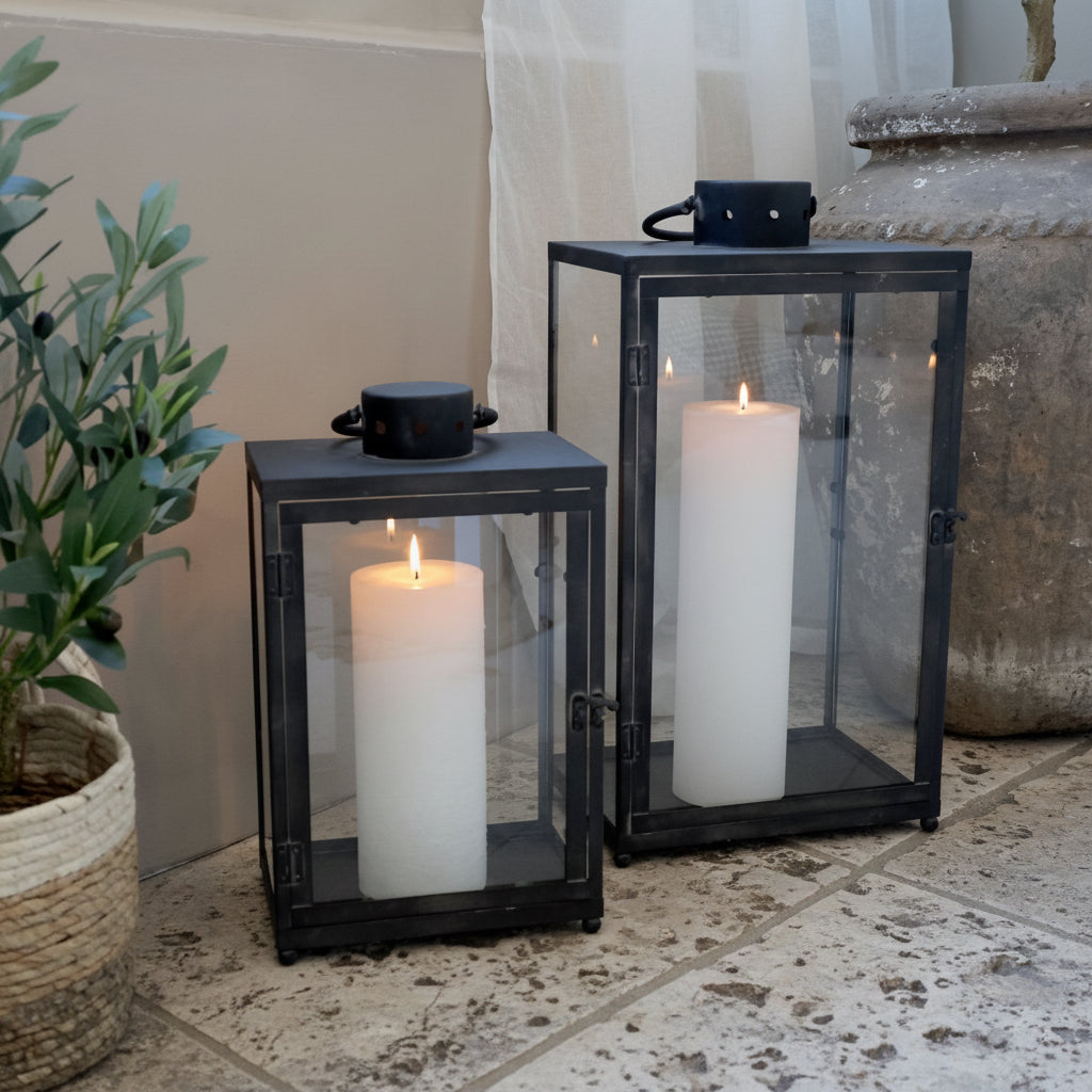 Two black lanterns with white candles inside on a light wooden floor.