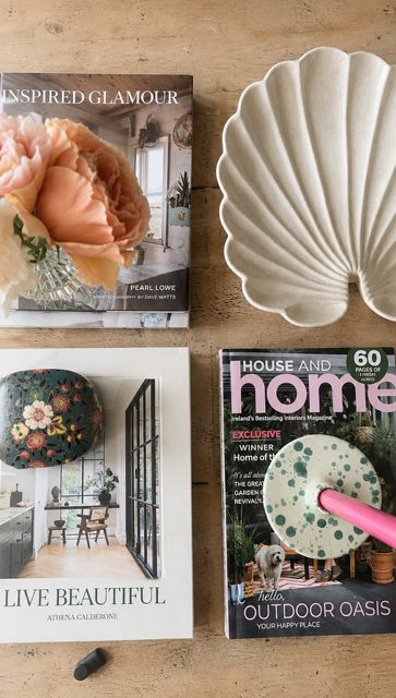A styled flat-lay on a light wood surface featuring a white scalloped shell-shaped ceramic dish, three magazines (Inspired Glamour, Live Beautiful, and House and Home), and a small black accessory.
