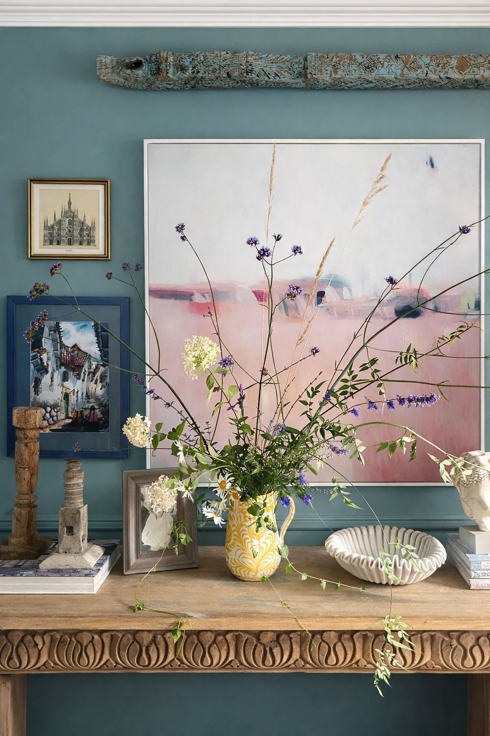 Decorative console with wild flowers, a yellow painted Spanish vase, and framed pictures on a wooden console table against a teal wall.