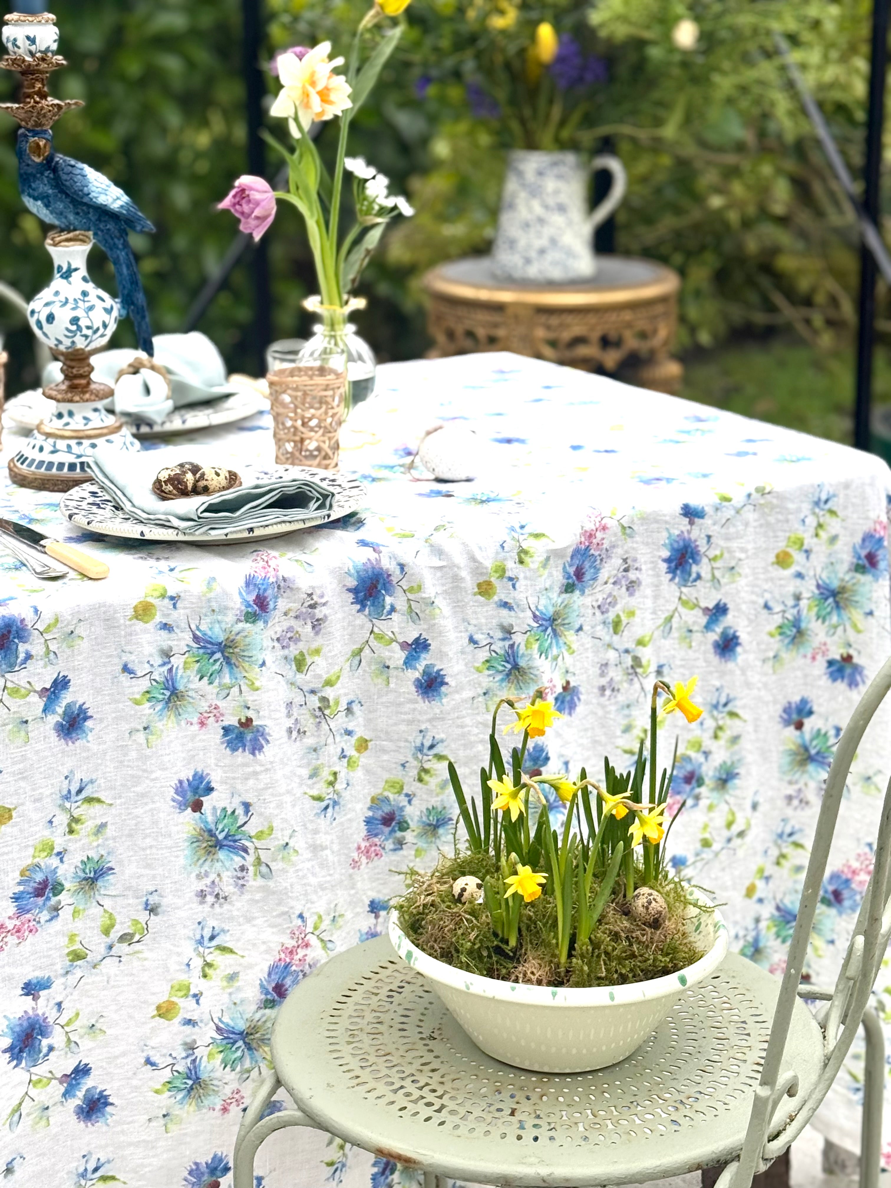 Outdoor table setting with floral tablecloth, plants, and decorative items.