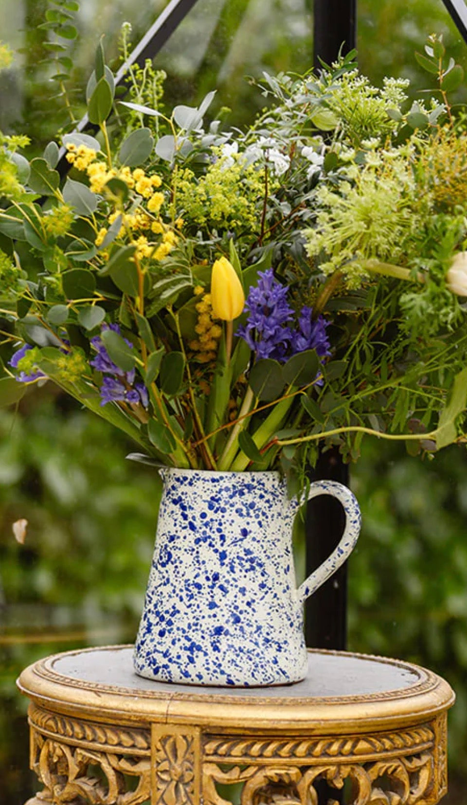 Floral arrangement in a blue and white speckled pitcher on a decorative table outdoors.