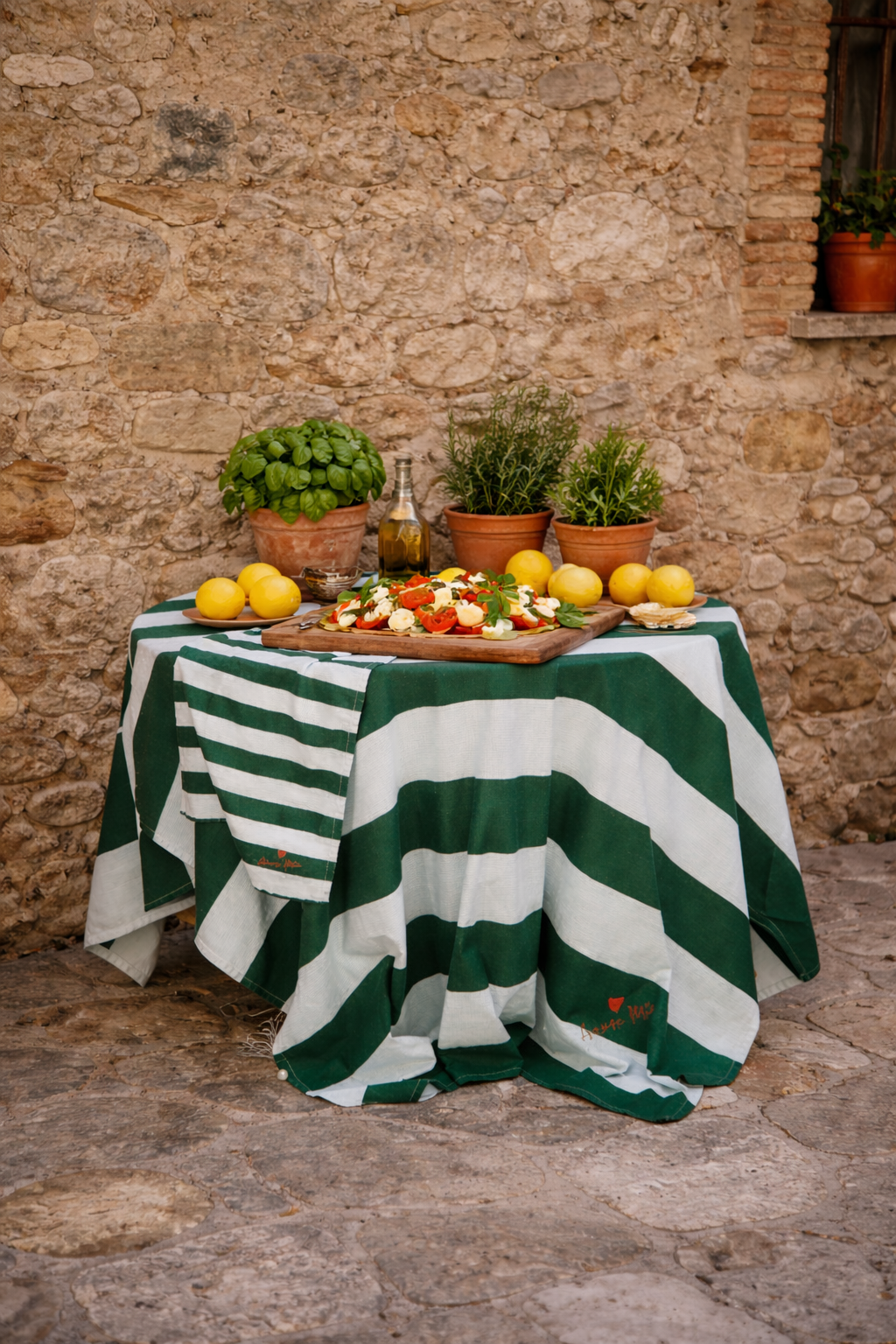 Table with a light blue an green striped tablecloth, loose lemons and potted herbs against an old stone wall.