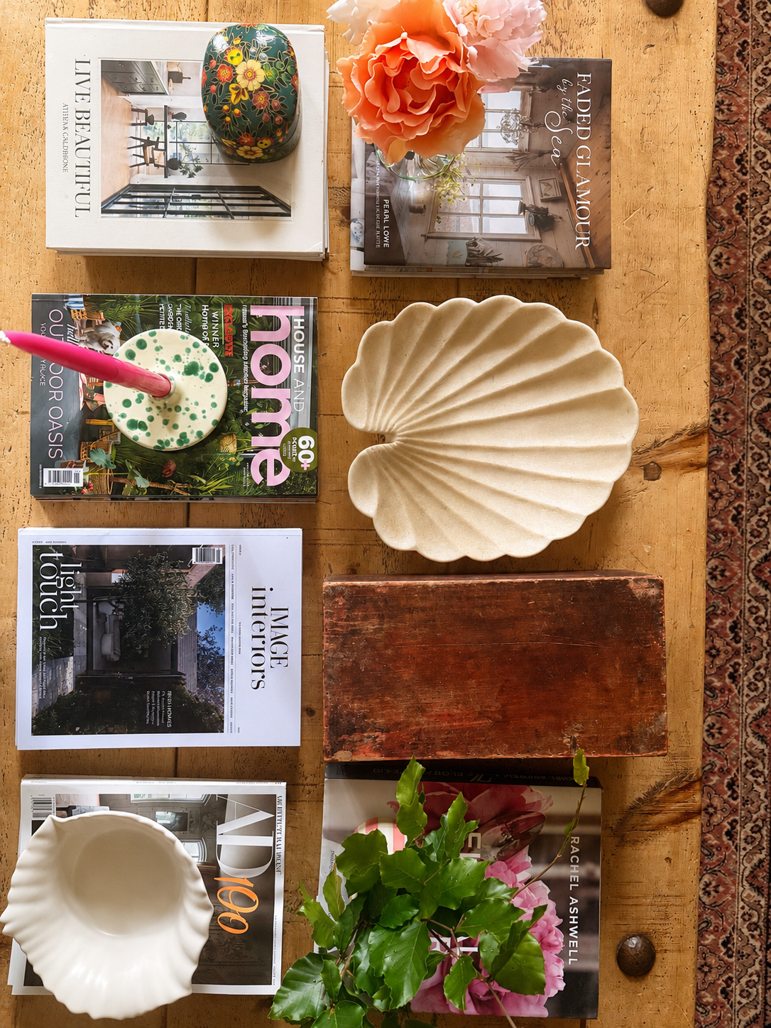 A styled flat-lay arrangement on a rustic wood coffee table featuring a marble shell plate, green splatter ceramic candleholder, a rectangular wooden box, stacks of magazines and coffee table books, apricot and pink coloured roses, and a cerise pink candle.