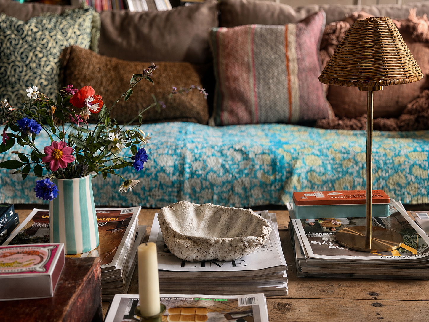 A cozy, rustic living room scene featuring a wooden coffee table styled with a striped ceramic vase of flowers, a textured stone bowl, a candle, and a brass lamp, set against a background of a patterned blue and yellow sofa with decorative pillows.