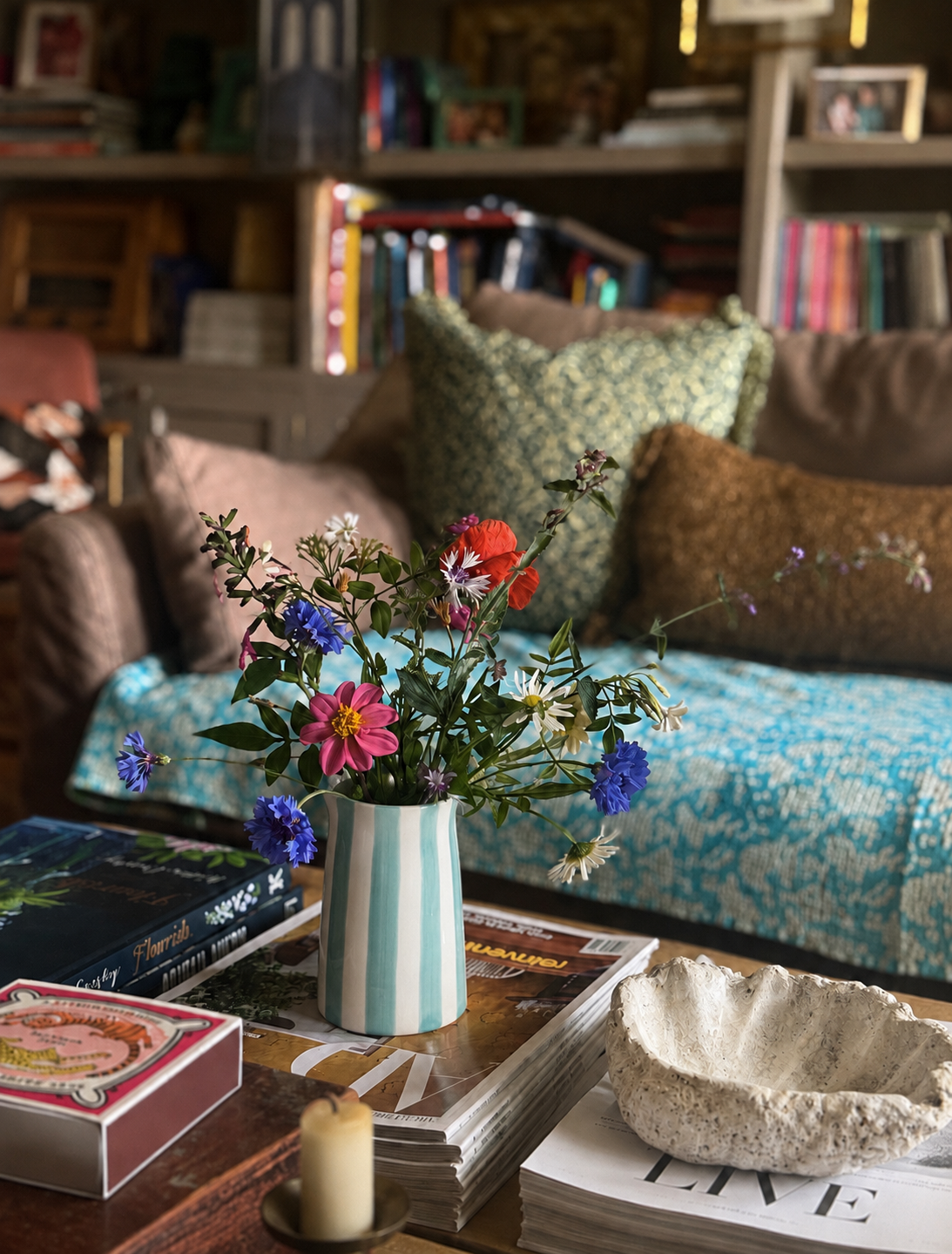 A turquoise striped vase of mixed wildflowers sits on a table in a cozy, book-filled living room with a colourful sofa in the background.