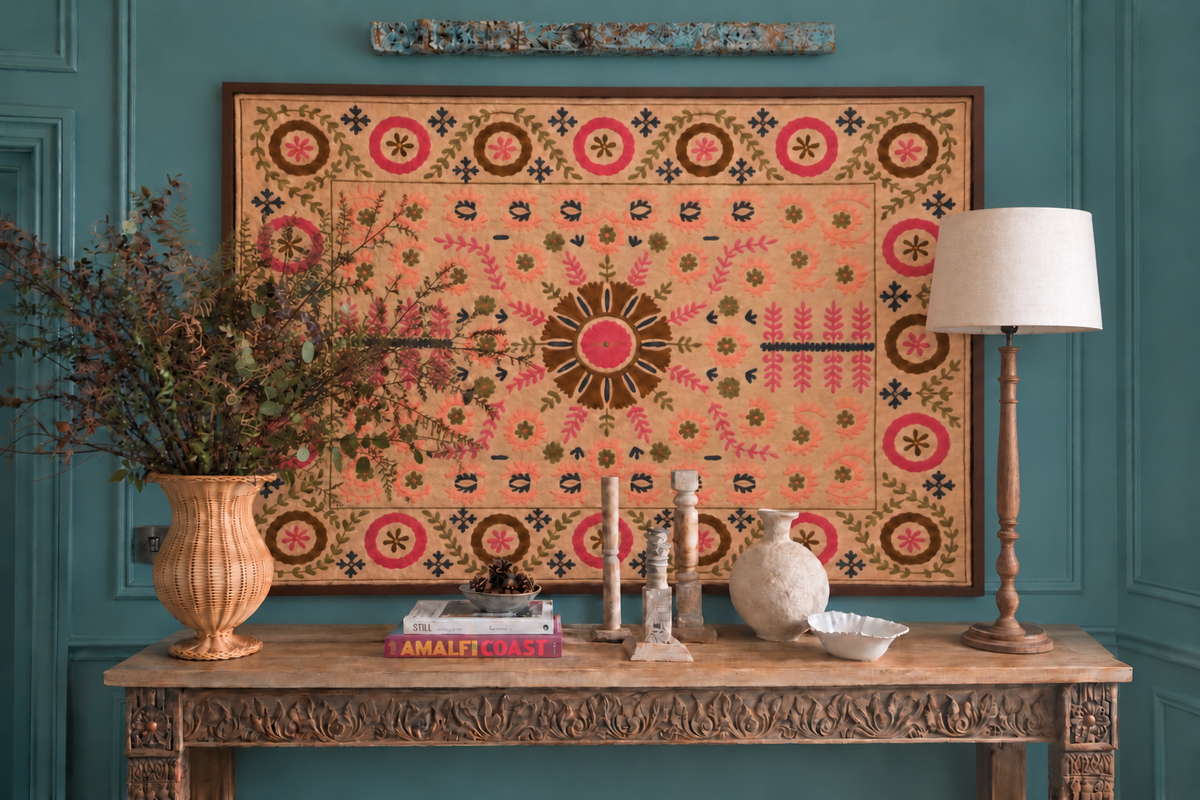 An eclectic scene with a large framed suzani textile hung on a teal panelled wall over a carved Indian console table decorated with a tall lamp with cream shade, books, a large rattan urn filled with bracken and branches; a cluster of wooden candle holders , a papier-mache vase and a white bowl.