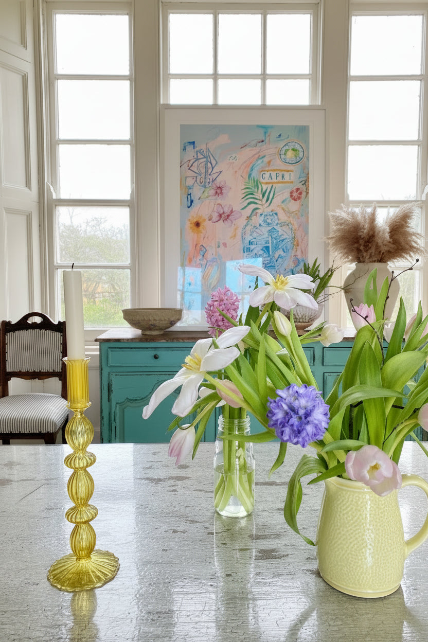 Yellow glass candle holder on rustic white table beside yellow jug of colourful flowers with  large window and colourful art in background