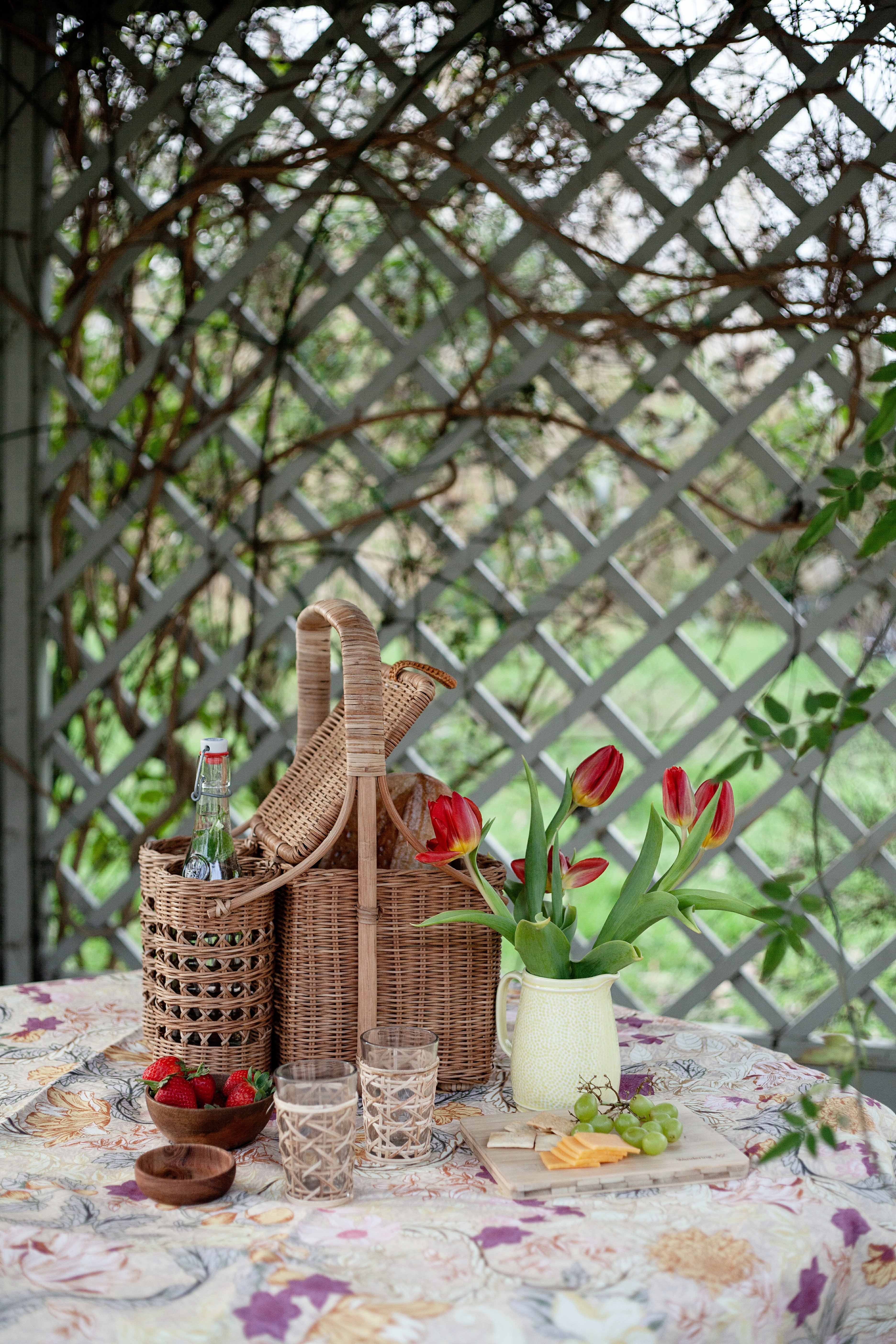 Outdoor setting with a picnic basket, flowers, and drinks on a table with a lattice background.