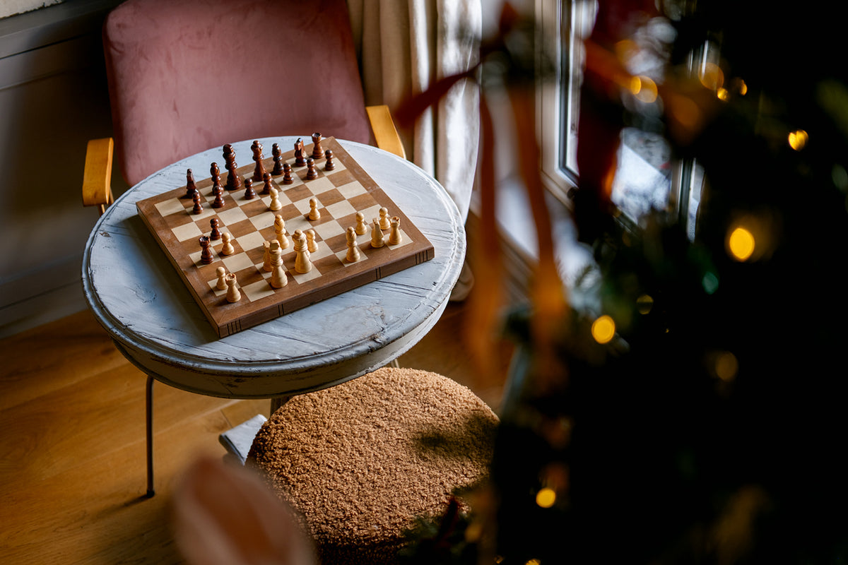 A wooden chessboard with dark and light chess pieces sits on a round white table in a cozy indoor setting with a pink armchair, a fuzzy brown pouf, and a blurred Christmas tree with warm lights in the foreground.