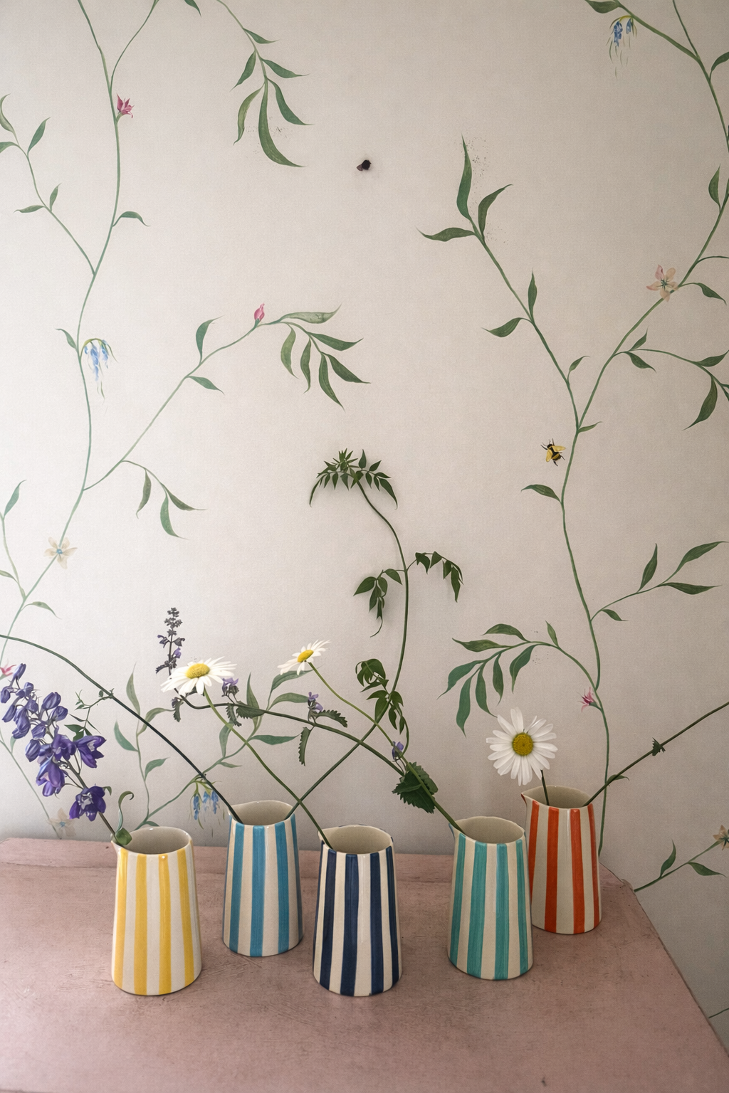 Five colorful striped vases with flowers against a wall with leaf patterns