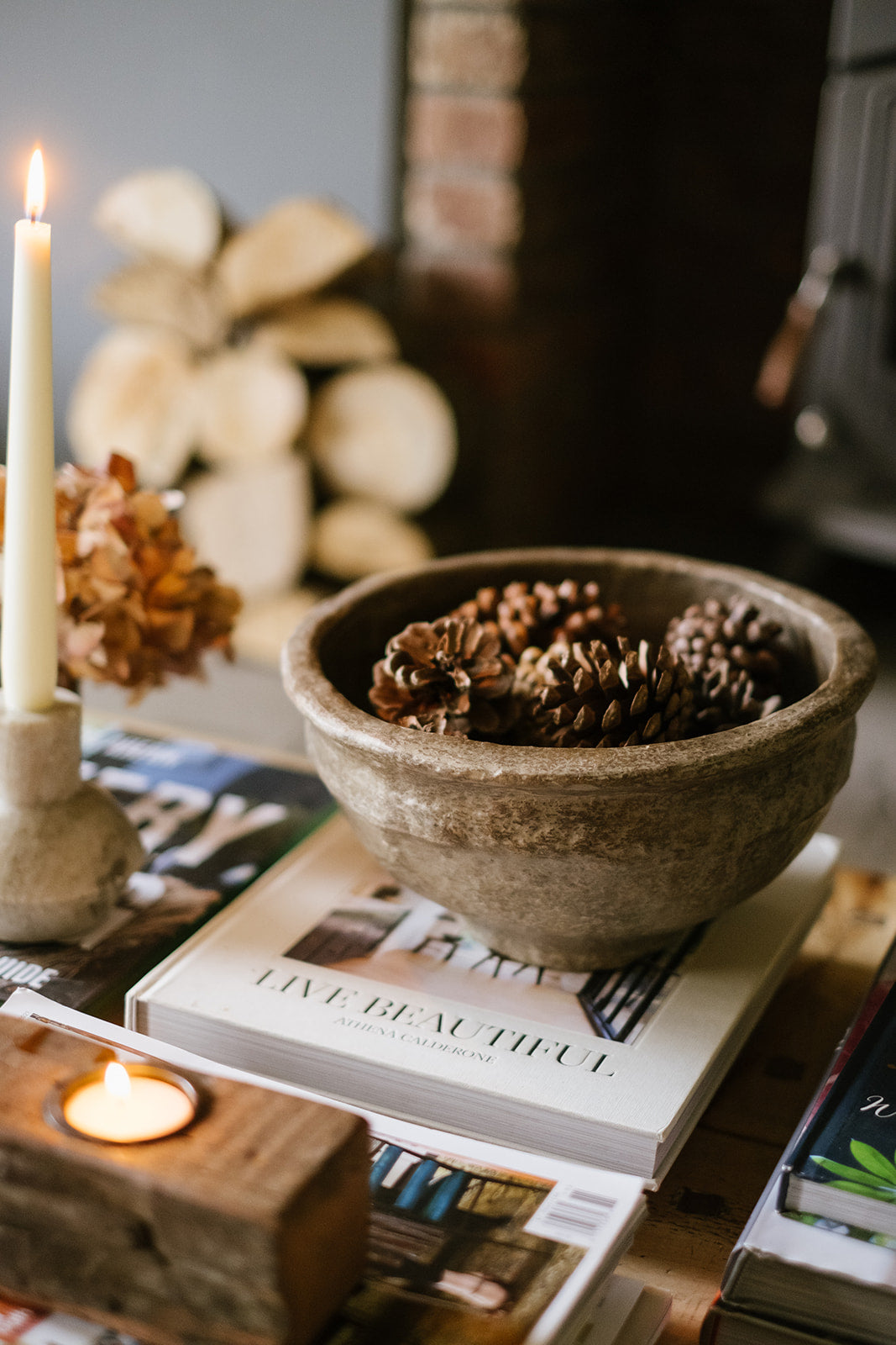 Grey textured papier-mache Bowl with pinecones on a stack of books with candles and decorative items.