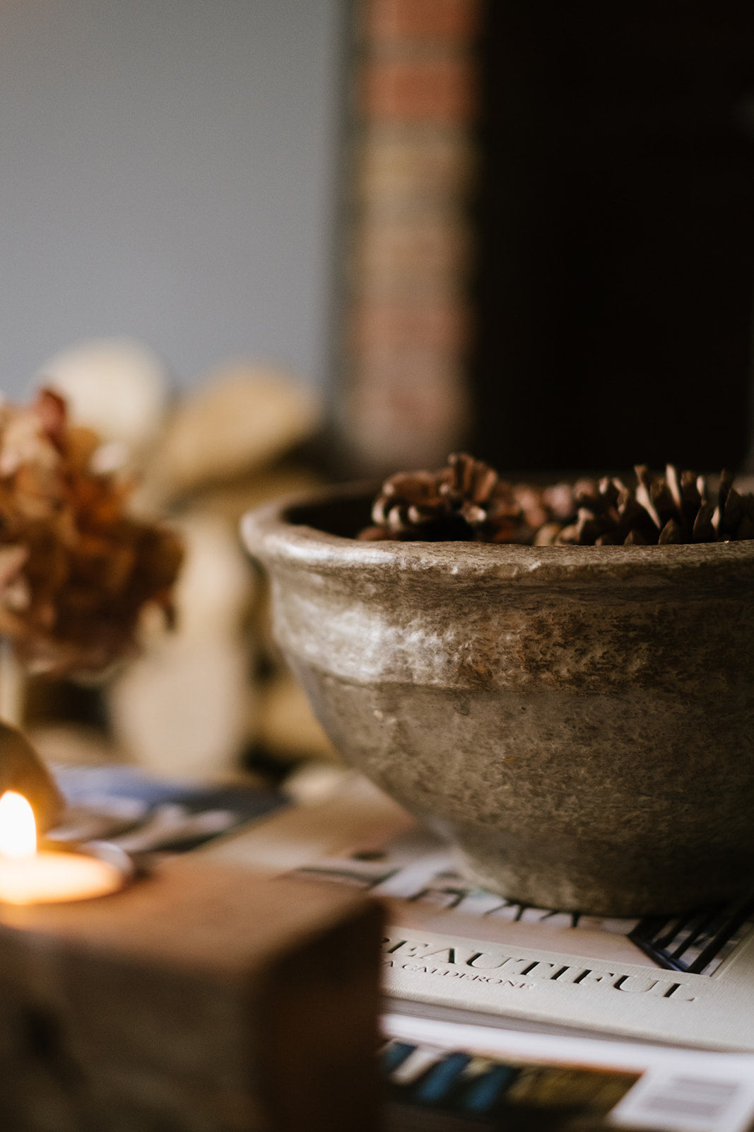 Grey textured papier-mache Bowl with pinecones on a stack of books with candles and decorative items.