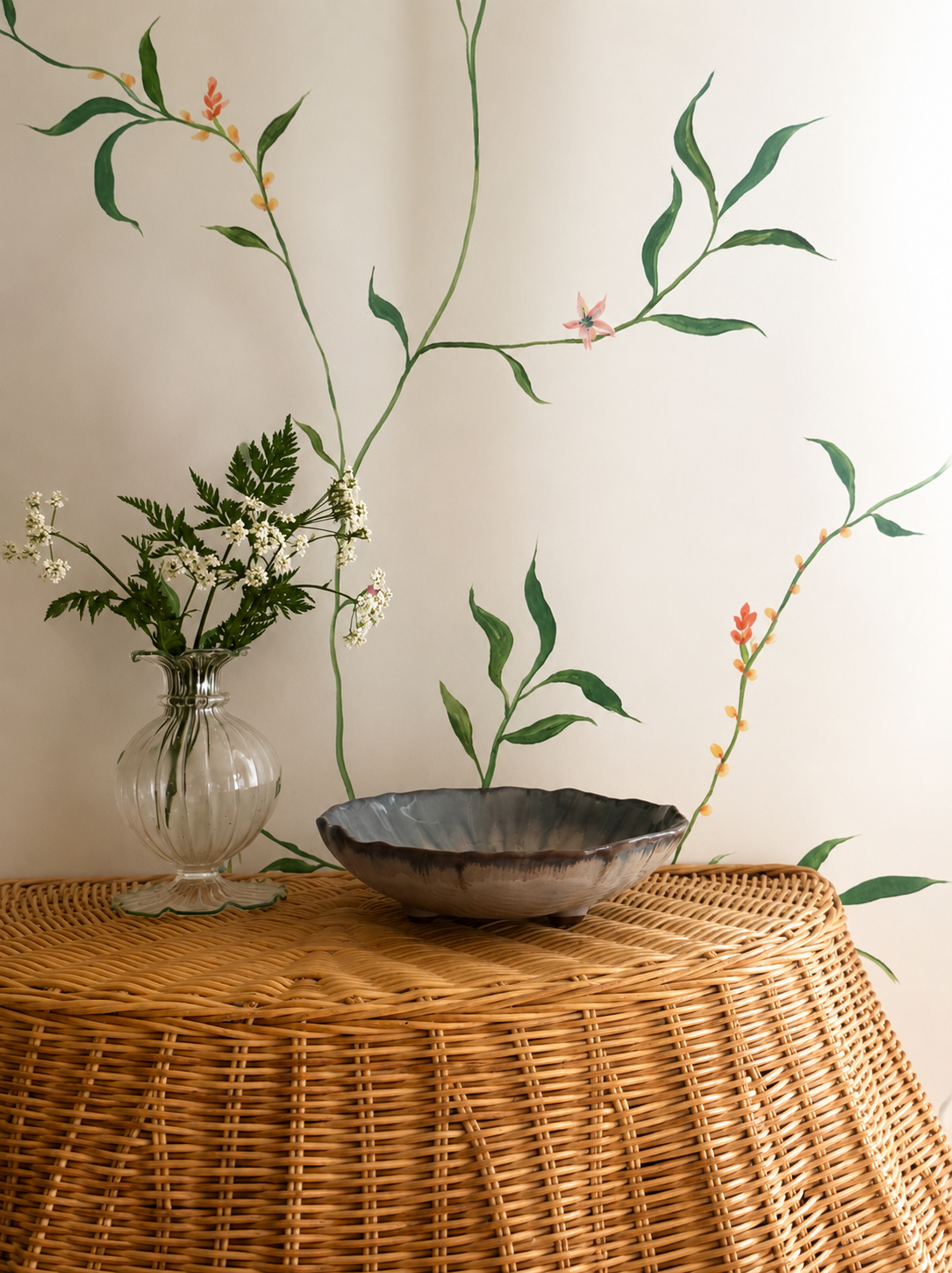 Rattan skirted console table with small footed glazed bowl and vase of green flowers against hand painted mural