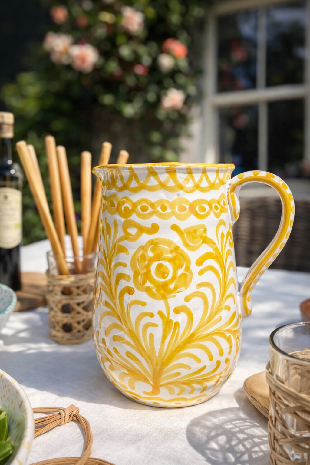 Close up of handmade Spanish pitcher in yellow traditional motif in a sunny outdoor setting with breadsticks and apricot climbing roses in the background