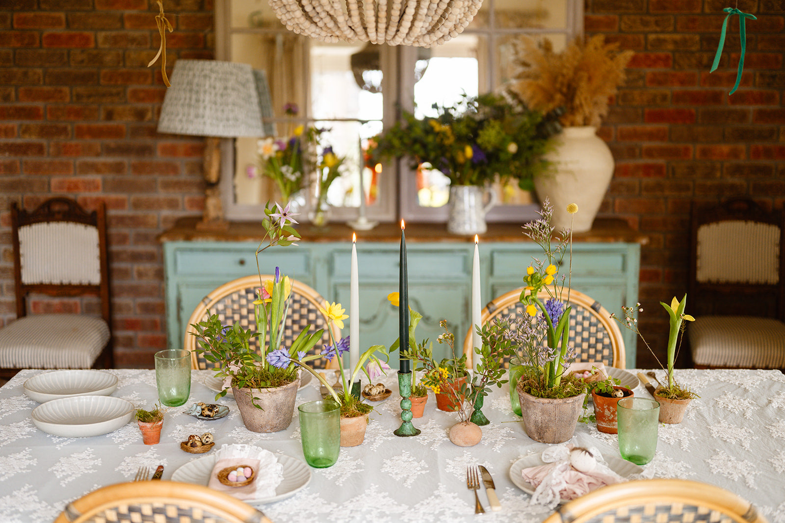 Romantic garden table setting with lace tablecloth, woven bistro chairs, terracotta pots, spring bulbs and flowers, verdigris candle holders with green candles against a rustic brick wall and turquoise cabinet