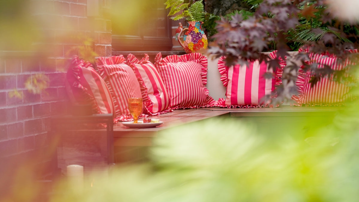rBright red and peach striped ruffled cushions arranged on a wooden bench in an outdoor patio setting, framed by soft green and pink foreground foliage.